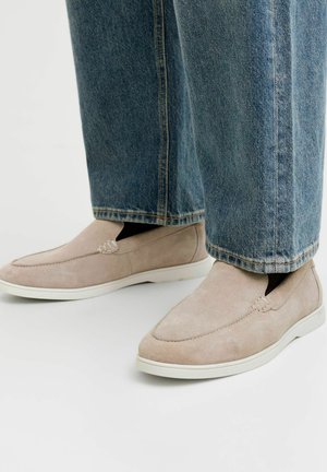 Beige ruskind slip-on sko båret med blå denim jeans, vist på hvid baggrund med synlige syninger og tekstureret stof.