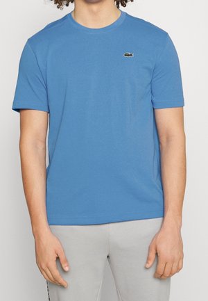 Homme portant un t-shirt Lacoste bleu à manches courtes et un pantalon gris clair, debout devant un fond blanc uni.