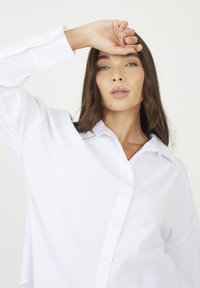 Donna con i capelli lunghi e castani e occhi verdi, che indossa una camicia bianca a pulsante larga, alzando la mano sinistra vicino alla fronte.