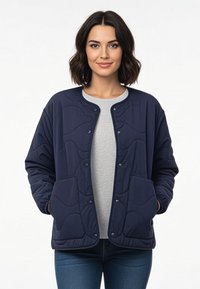 Frau mit schulterlangem dunklem Haar, die eine marineblaue gesteppte Jacke über einem grauen Shirt trägt, die Hände in den Taschen, stehend vor einem weißen Hintergrund.