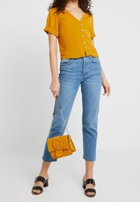 Sininen lyhythihainen pusero, jossa on V-pääntie ja nappiyksityiskohta, yhdistettynä vaaleansinihin pussihousuihin ja keltaiseen mokkanahkaiseen viestin laukkuun. Mustat liukuva kengät.