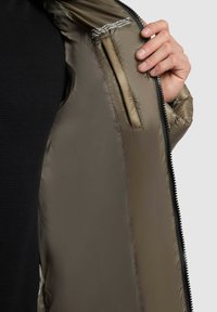 Olive green puffer jacket interior with a smooth, shiny texture. Includes a zippered pocket and printed label near the neckline.