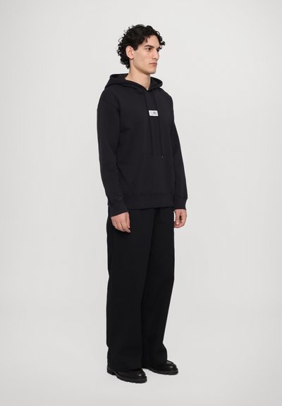 Sweatshirt à capuche noir avec une poche kangourou à l'avant, arborant un petit écusson blanc. Porté avec un pantalon noir ample et des bottes.