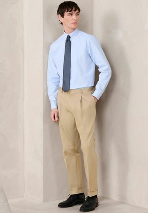 Mann trägt hellblaues Hemd, marineblaue Krawatte, beige Hose und schwarze Lederschuhe, steht vor einer beigen Wand und schaut nach rechts.