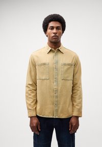 Cămașă din denim bej cu închidere cu butoni, două buzunare pe piept și guler deschis, având o suprafață ușor texturată și cusături vizibile.