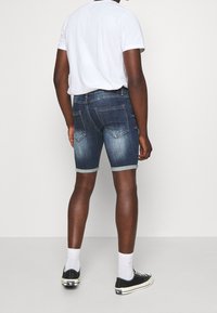 Bermudas de mezclilla con un lavado azul oscuro, dobladillo vuelto, diseño de cinco bolsillos y detalles de costura visibles, combinadas con una camiseta blanca lisa y zapatillas.