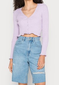 Donna che indossa un cardigan a coste lavanda, corto e con bottoni, abbinato a pantaloni corti in denim strappati, leggeri e a vita alta di colore blu.