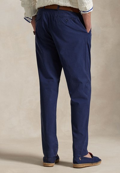 Muškarac odjeven u mornarski plave hlače chino, smeđi remen, krem džemper i mornarske espadrile s pletenim potplatima, stoji s rukama u džepovima.