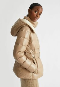 Giacca a vento beige con cappuccio, cuciture dettagliate, vita con cintura e tasche laterali. Texture lucida con un colletto bicolore.