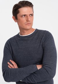Dunkelgrauer strukturierter Pullover mit langen Ärmeln und einem weißen Rundhalsausschnitt. Das Model hat die Arme verschränkt und schaut zur Seite.