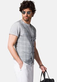 Szary kamizelka w kratę z niebieskimi akcentami, krótkie rękawy i guziki; zestawiona z szarym t-shirtem i białymi spodniami, trzyma czarną torbę.