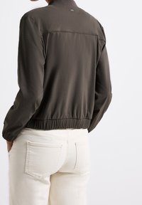 Mörkgrön bomberjacka med ribbad krage och elastisk midja, med långa ärmar och slät yta. Bärs med vita jeans.