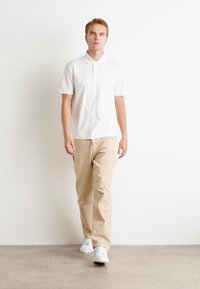 Camisa polo branca com colarinho, combinada com calças beje claras e ténis brancos, de pé contra um fundo branco liso.