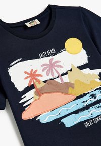 Camiseta de algodón azul marino con una escena de playa multicolor que incluye palmeras, olas y un sol. El texto dice "PLAYA SALDA" y "GRAN VERANO."