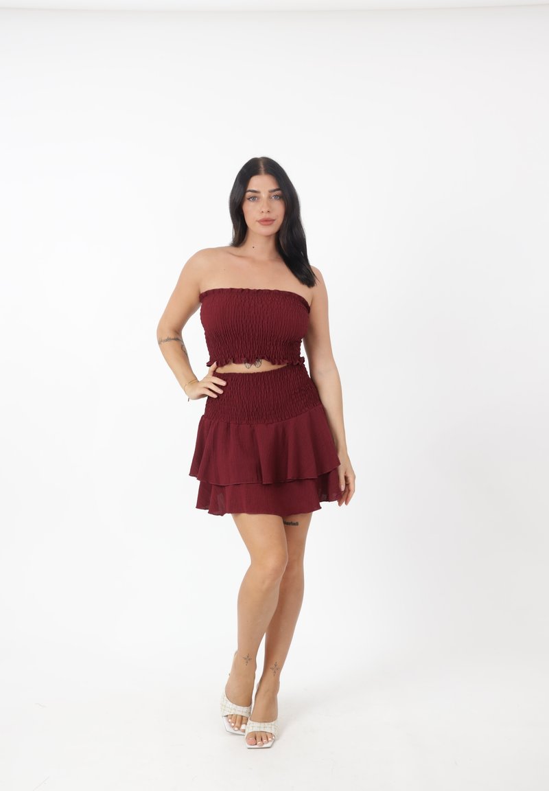 Burgunderfarbendes Zweiteiler-Kleid mit einem gerafften trägerlosen Oberteil und mehrlagigem Rock. Das Material wirkt strukturiert. Hellbeige Sandalen ergänzen das Outfit.
