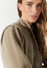 Lichtbruine cardigan gemaakt van zachte gebreide stof. Kenmerkt zich door een geribde kraag, pouf-mouwen en decoratieve knoopdetails aan de voorkant.