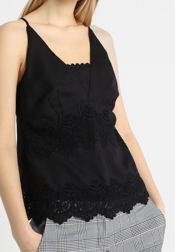 Femme portant un haut noir sans manches avec des détails en dentelle et un pantalon à carreaux gris, mains dans les poches, fond clair uni.