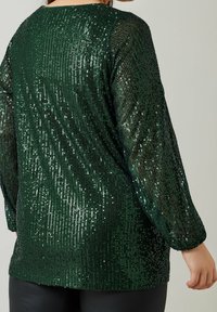 Femme portant un haut à manches longues vert foncé recouvert de sequins scintillants, vue de dos sur un fond uni.