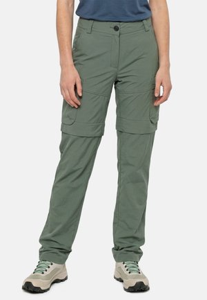 Persoon draagt olijfgroene afritsbare cargobroek en beige wandelschoenen met groene veters, staand tegen een witte achtergrond.