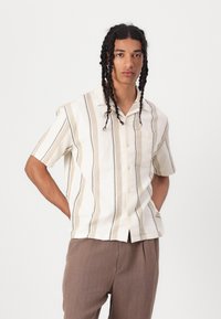 Chemise à manches courtes, boutonnée, de couleur crème avec des rayures verticales beiges et marine. Tissu léger avec une poche sur le côté gauche.