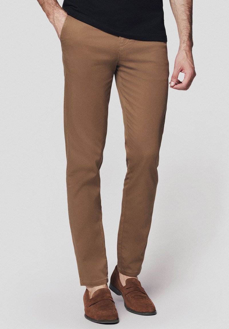 Chinos wąskie brązowe o gładkiej teksturze, z szlufkami na pasek i bocznymi kieszeniami, w połączeniu z brązowymi loafersami. Tkanina wydaje się lekka i trwała.