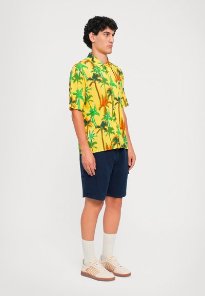 Kurzärmliges gelbes Hemd mit grünem und orangefarbenem Palmenmuster, kombiniert mit marineblauen Shorts, hellen Socken und beigen Sneakers mit Streifen.