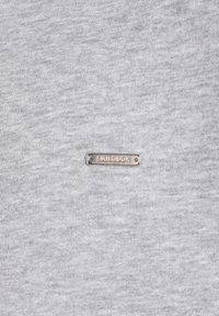 T-shirt en coton gris avec une petite étiquette métallique portant un texte gravé, centrée à l'avant, avec une texture lisse et uniforme.