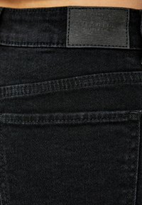 Närbild av svarta denimjeans bakficka med sömnadsdetaljer och en svart läderlogg på midjebandet.