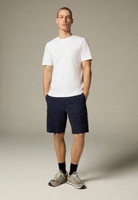 Tricou alb cu mânecă scurtă, şorturi cargo marine cu buzunare și adidași gri cu șosete negre. Modelul pozează cu mâinile în buzunare.