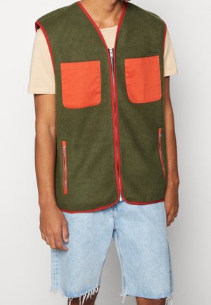 Homme portant un gilet sans manches vert olive avec des poches et bordures orange, un t-shirt beige et un short en jean bleu clair effiloché.