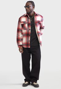 Rot-schwarz kariertes Hemd-Jacke mit zwei Brusttaschen; über einem schwarzen T-Shirt getragen. Schwarze, weite Hose und Sonnenbrille vervollständigen den Look.