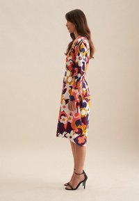 Musterkleid mit lebendigen Blumenmotiven in Pink, Gelb und Marineblau, mit langen Ärmeln, einem taillierten Gürtel und Midi-Länge. Getragen mit schwarzen hohen Schuhen.