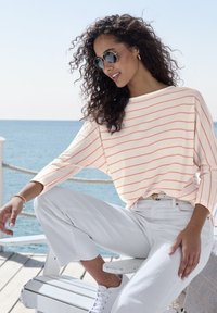 Leichter Strickpullover mit korallenfarbenen Streifen, entspannte Passform, weite Ärmel. Kombiniert mit weißen, knöchellangen Hosen und weißen Sneakers, gestylt mit Sonnenbrille.
