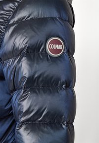 Manga de chaqueta acolchada azul marino oscuro con un parche redondo del logotipo de la marca Colmar en burdeos y blanco.