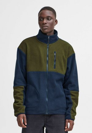 Fleecejacke mit hohem Kragen, in grünen und marineblauen Farbblöcken, ausgestattet mit einer Vorderreißverschlusstasche und Seitentaschen. Glatte Textur.