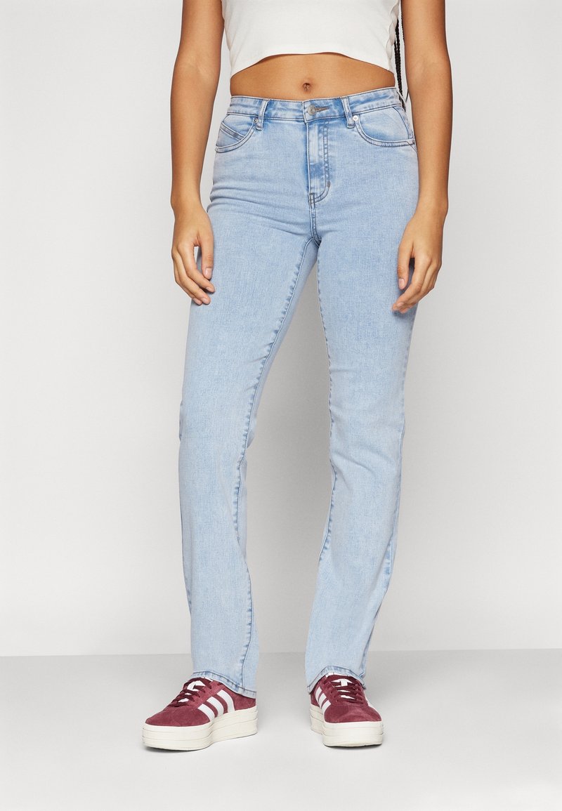 Femme portant un jean skinny taille haute bleu clair, un top court blanc et des baskets bordeaux avec des bandes blanches, debout devant un fond uni.