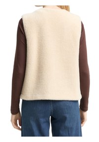Personne portant un gilet duveteux de couleur crème par-dessus une chemise marron manches longues et un jean bleu, vue de dos.