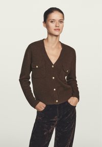 Cardigan in lana marrone con scollo a V profondo, dotato di due tasche anteriori e bottoni dorati. Abbinato a pantaloni scuri e strutturati.