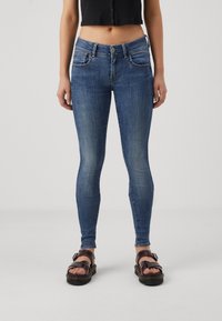 Jean skinny bleu en denim avec un lavage décoloré, comportant cinq poches, des passants de ceinture et une fermeture à bouton. Porté avec des sandales compensées noires.