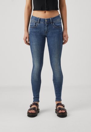 Jean skinny bleu en denim avec un lavage décoloré, comportant cinq poches, des passants de ceinture et une fermeture à bouton. Porté avec des sandales compensées noires.