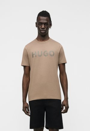 Muž v béžovom tričku s krátkym rukávom s nápisom "HUGO" a čiernymi šortkami po kolená stojí pred jednoduchým bielym pozadím.