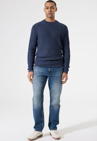 Marineblauwe ribgebreide trui met een ronde hals, gecombineerd met vervaagde blauwe recht afgesneden jeans en witte sneakers op een effen achtergrond.