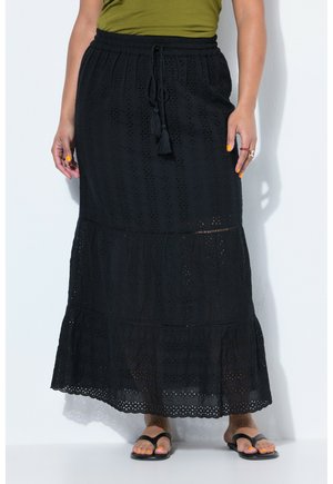 Frau trägt einen schwarzen, knöchellangen Rock mit Lochmuster und Quasten-Zugband, kombiniert mit schwarzen Sandalen und einem olivgrünen Oberteil.