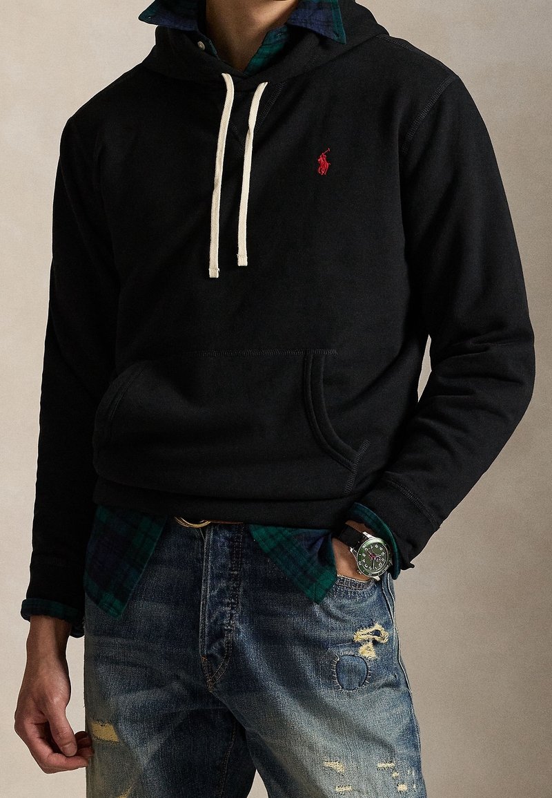 Personne portant un sweat à capuche noir avec des cordons blancs, une chemise à carreaux verte en dessous, un jean bleu déchiré et une montre-bracelet au cadran vert.