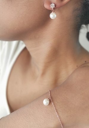 Boucles d'oreilles en forme de goutte de perle avec des accents en or rose ; elles présentent un petit cristal sur le clou, une perle blanche lisse et un bracelet délicat en chaîne avec une perle assortie.