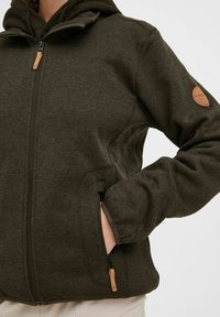 Temno zelena zip-up jakna z teksturirano površino, ki ima kapuco, stranske žepke in usnjen logotip na levem rokavu.
