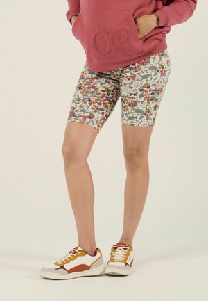 Personne portant un short à motifs floraux mi-cuisses, un sweat-shirt rouge rouille avec les mains dans la poche avant, et des baskets blanches avec des accents orange.