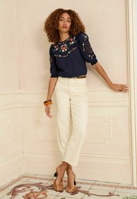 Navy blouse with floral embroidery, puffed sleeves; cream straight-leg trousers; tan heeled sandals; wooden bangle accessory.