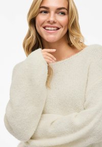 Uśmiechająca się kobieta z blond włosami, ubrana w miękką, kremową sweter z puszystej tkaniny, z długimi, luźnymi rękawami, na białym tle.