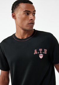 Czarny t-shirt sportowy z okrągłym dekoltem, kontrastowym czerwonym szwem i logo z napisem "A.T.H" oraz "ATHLETIC RUN THE RACE" w białym i czerwonym kolorze.